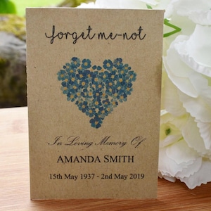 May include: A brown paper packet of forget-me-not seeds with a heart-shaped design of blue flowers. The packet reads "forget me-not" and "In Loving Memory Of AMANDA SMITH 15th May 1937 - 2nd May 2019".