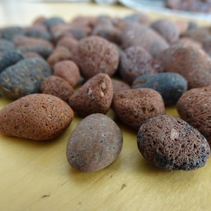 Può includere: Una collezione di piccole rocce vulcaniche porose in tonalità marrone rossastro e grigio antracite scuro. Queste pietre presentano una consistenza ruvida e bucherellata tipica della roccia lavica, con forme e dimensioni irregolari varie.