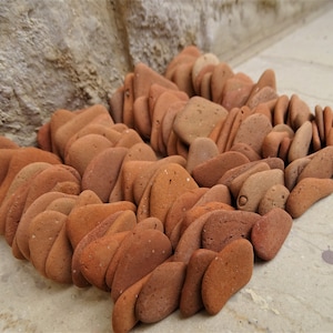 May include: A collection of numerous small, flat, terracotta-colored stones arranged in a dense, overlapping cluster. Each stone features a smooth, rounded shape with a matte finish, displaying various shades of earthy reddish-brown tones.