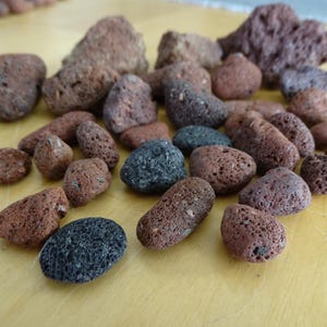 May include: A collection of small, porous volcanic lava rocks in various shades of reddish-brown, dark gray, and black. Each stone features a rough, pitted texture with visible air pockets, scattered across the surface in an irregular arrangement.