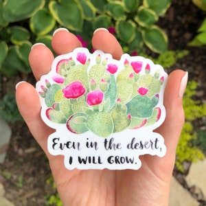 Może przedstawiać: Naklejka z akwarelową ilustracją kaktusa z r&oacute;żowymi kwiatami. Naklejka zawiera napis "Even in the desert, I WILL GROW" czarnym pismem. Kaktus jest zielony z czerwonymi akcentami.