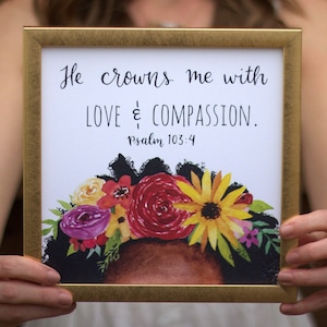 May include: A framed print with a white background and gold frame. The print features a watercolor illustration of a woman with a crown of flowers on her head. The text reads "He crowns me with LOVE & COMPASSION. Psalm 103:4".