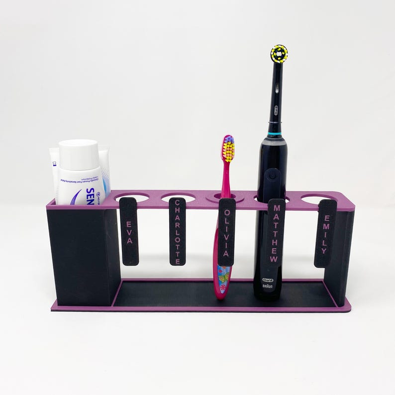 May include: A black and purple toothbrush holder with five slots. The holder has labels for each slot: Eva, Charlotte, Olivia, Matthew, and Emily. There is a pink toothbrush in the Olivia slot and a black electric toothbrush in the Matthew slot. A tube of toothpaste is in the Eva slot.