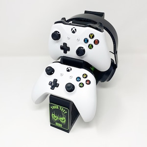 Op de afbeelding: Een zwarte standaard met een groene en zwarte afbeelding met de tekst "YOUR TEXT HERE" houdt twee witte Xbox-controllers en een zwarte headset vast.