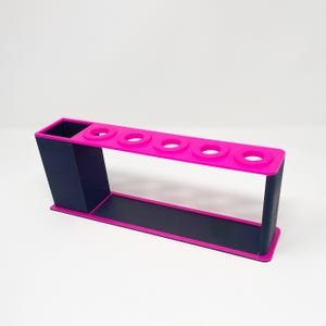 May include: A pink and black plastic organizer with five slots for holding pens or pencils. The organizer has a separate compartment for storing other items.