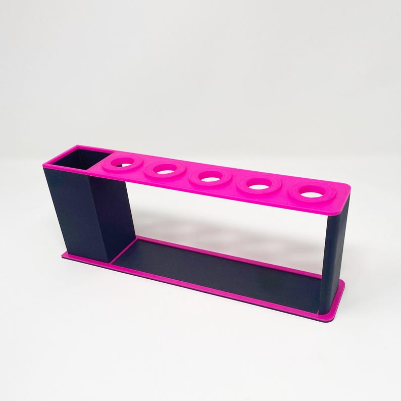 May include: A pink and black plastic organizer with five slots for holding pens or pencils. The organizer has a separate compartment for storing other items.