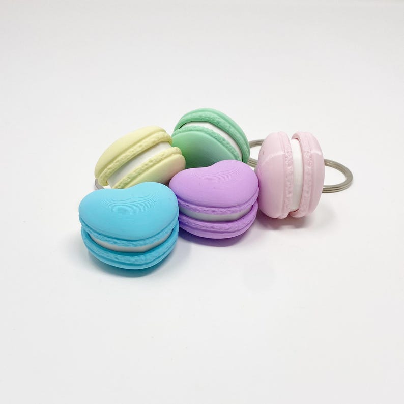 Op de afbeelding: Een set van vijf pastelkleurige macaron sleutelhangers. De sleutelhangers zijn gevormd als traditionele macarons, met een witte vulling en een lichtgroene, gele, lichtblauwe en paarse buitenschaal.