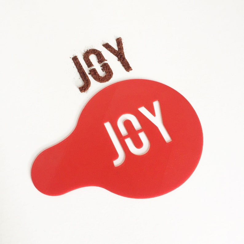 May include: Red plastic stencil with the word "JOY" cut out in white. The stencil is on a white surface with brown powder sprinkled on the surface, forming the word "JOY".