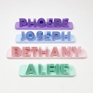 May include: Four personalised name blocks with names spelled out in a retro style font. The names are Phoebe, Joseph, Bethany, and Alfie. The blocks are in different colours: purple, blue, pink, and green.