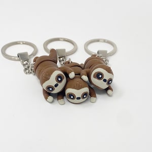 Puede incluir: Tres llaveros de perezoso marrones con anillos de llave plateados. Los perezosos están colgando boca abajo con los brazos y las piernas extendidos.