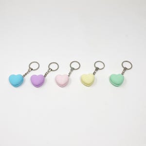 Op de afbeelding: Vijf hartvormige sleutelhangers in verschillende pastelkleuren: lichtblauw, paars, roze, geel en groen. Elke sleutelhanger heeft een hartvormige hanger die lijkt op een macaron.
