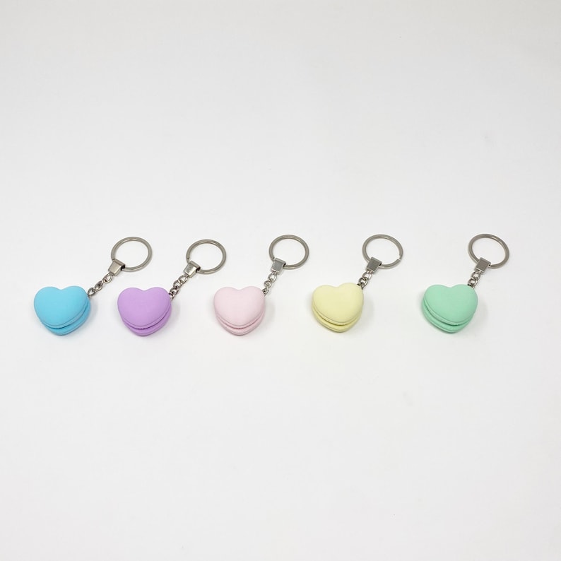 Op de afbeelding: Vijf hartvormige sleutelhangers in verschillende pastelkleuren: lichtblauw, paars, roze, geel en groen. Elke sleutelhanger heeft een hartvormige hanger die lijkt op een macaron.