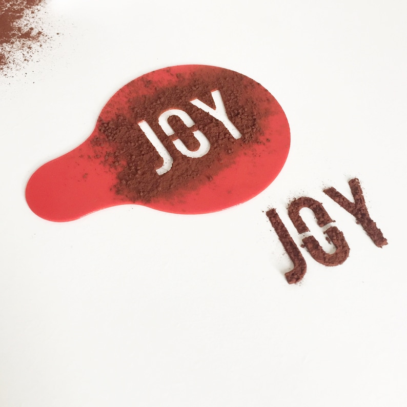 May include: Red plastic stencil with the word "JOY" cut out. The stencil is on a white surface with cocoa powder dusted over it, creating the word "JOY" in the powder.