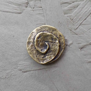 Könnte beinhalten: Ein bronzefarbener, kreisförmiger Anhänger mit einem erhabenen Spiraldesign. Der Anhänger hat eine strukturierte Oberfläche und ein rustikales, antikes Aussehen. Die Spirale ist innerhalb des Kreises zentriert und bildet einen Brennpunkt.