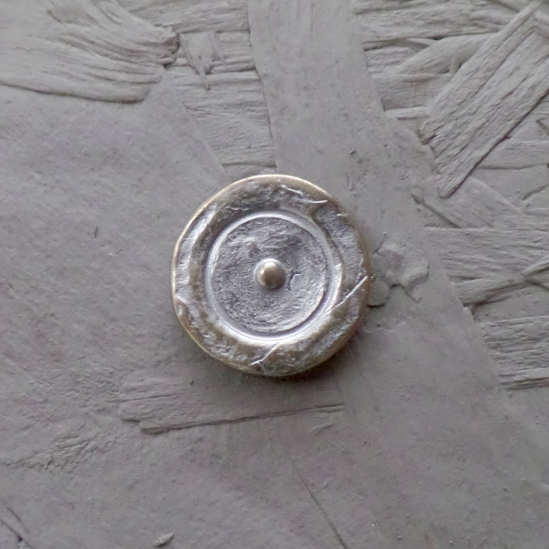 May include: A silver-coloured metal button with a concentric circle design and a small raised dot in the centre. The button is attached to a grey textured surface.