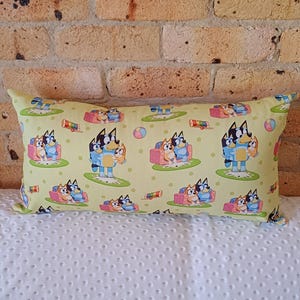 May include: A yellow and green rectangular cushion with a repeating pattern of blue dogs with orange ears. The dogs are sitting on a sofa, standing, and holding a ball. There are also some toys on the cushion, including a xylophone and a ball.