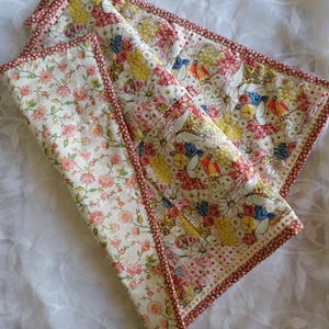 May include: A stack of floral fabric napkins with a red dotted border. The napkins feature a variety of colourful flowers on a cream background. One napkin has a pink floral pattern.