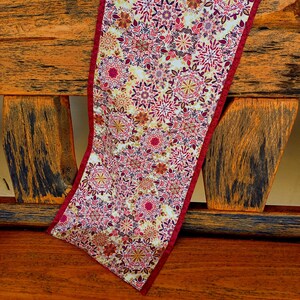 May include: A decorative table runner featuring a floral design in red, pink, and gold hues against a cream backdrop. The runner has a solid red border and is displayed on a wooden surface.