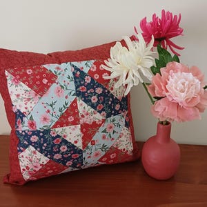 May include: A patchwork cushion with red, blue, and white floral patterns. The cushion has a solid red border. A vase with pink and white flowers sits beside it, adding a touch of colour.