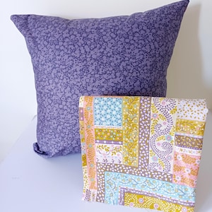 May include: A purple cushion with a floral pattern and a folded fabric with a patchwork design. The fabric features squares and rectangles with floral and geometric patterns in shades of pink, blue, and yellow.