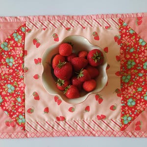 May include: A strawberry-themed placemat featuring a central bowl of fresh strawberries. The placemat has a light pink base with a strawberry and floral pattern. Red and white striped borders and floral triangles frame the central section. The placemat is approximately 40 cm long.