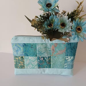 May include: A teal and turquoise patchwork cosmetic bag with a light blue zip and trim. The bag is decorated with artificial blue flowers with brown centres and green leaves. The bag is made of quilted fabric.