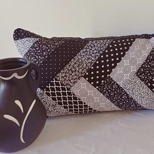 May include: A black and white patchwork cushion with a variety of patterns, and a dark brown vase with a white floral design. The cushion's patterns include dots, floral prints, and geometric shapes. The vase is a decorative item.
