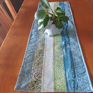 May include: A rectangular table runner with a patchwork design in shades of blue, green, and white. A small white ceramic planter with a green plant sits on top. The runner is approximately 30 cm wide.