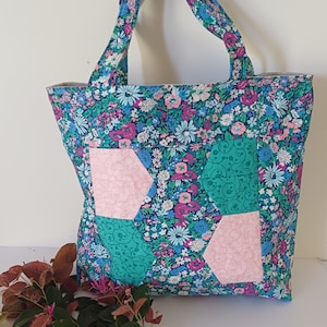 May include: A tote bag featuring a vibrant floral pattern in shades of blue, pink, and white. The bag includes two shoulder straps and is accented with two light pink and two teal geometric fabric patches stitched onto the front floral panel.