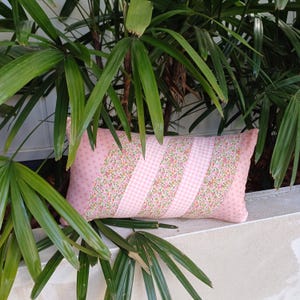 May include: A pink and white patterned lumbar cushion with a gingham and floral design. The cushion is sitting on a white surface.