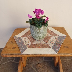 May include: A small wooden table with a floral patterned quilt and a potted cyclamen plant with bright pink flowers. The table has a natural wood finish and a rustic design, ideal for a cosy home decor.