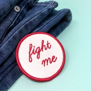 May include: A white embroidered patch with red stitching. The patch is round and says "fight me" in red cursive lettering. The patch is sitting on top of a stack of blue denim jeans.
