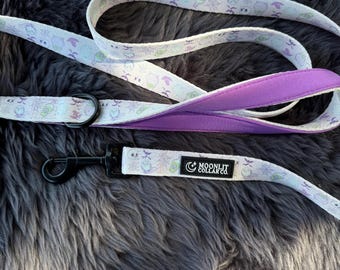 May include: A dog leash with a white background and a repeating pattern of Halloween-themed graphics in purple and green. The leash has a purple padded handle and a black metal clasp. The text "MOONLIT COLLAR CO." is printed on the leash.