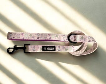 May include: A light pink dog leash with a black clasp and a matching handle. The leash features a repeating pattern of Halloween-themed graphics, including ghosts, spiderwebs, and potion bottles. The brand name "Moonlit Collar Co." is printed on the leash.