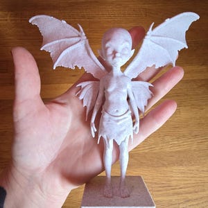 May include: White 3D printed figurine of a fairy with wings, standing on a square base. The fairy has a human-like body with wings and a small, delicate face.