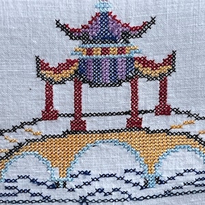 May include: A cross-stitch design of a pagoda with a bridge. The pagoda features a multi-colored roof in shades of purple, red, yellow, and blue. The bridge is yellow with blue accents, and the background is white.