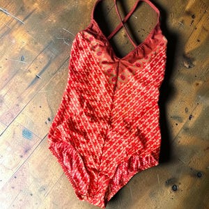 May include: A one-piece swimsuit in a vibrant red hue featuring a repeating white leaf-like pattern. The garment has a deep V-neckline with a decorative peacock feather-inspired border at the chest and thin, crisscrossing shoulder straps.