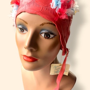 May include: A vintage red swim cap with a textured design and floral accents. The cap features a tag that reads "Aqua Fashions by US". The cap has a long red strap.