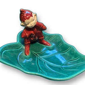 Könnte beinhalten: Eine Keramikfigur eines Elfen in roter Kleidung, der auf einer grünen, blattförmigen Schale sitzt. Der Elf hat ein weißes Gesicht und eine rote Zipfelmütze. Die Schale ist leuchtend grün mit Blattader-Details.