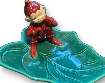 May include: A ceramic elf figurine in a red outfit, sitting on a green leaf-shaped dish. The elf has a white face and a red pointed hat. The dish is a vibrant green with leaf vein detailing.