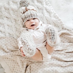 May include: A baby wearing a white knitted hat with a brown pom-pom and a brown inscription, and matching knitted booties. The baby is lying on a white knitted blanket.