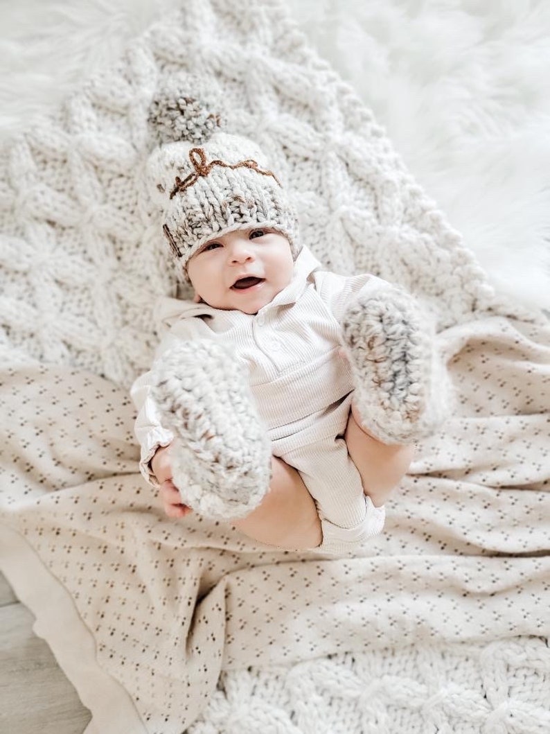 May include: A baby wearing a white knitted hat with a brown pom-pom and a brown inscription, and matching knitted booties. The baby is lying on a white knitted blanket.