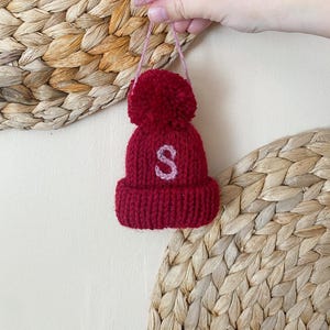 May include: A small, knitted red beanie with a matching pom-pom and a light pink embroidered "S" on the front. The beanie is suspended by a pink string. The background features woven textures.