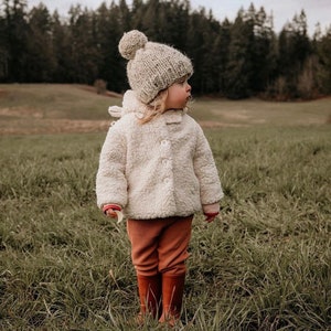May include: A young child wearing a cream-colored fuzzy jacket, brown pants, and brown rain boots. The child is wearing a light brown knit hat with a pom-pom on top. The child is standing in a field of green grass.