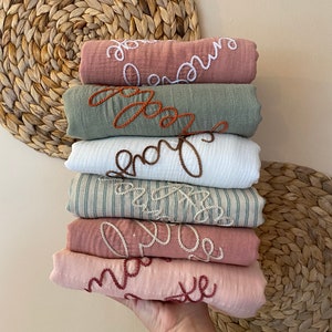 May include: A stack of seven rolled muslin blankets in various colors, including pink, green, and white. Each blanket features embroidered cursive text in contrasting colors, spelling out words like "love" and "dream". The blankets are held in a hand.
