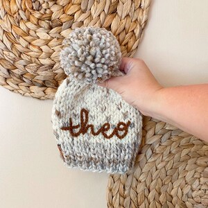 May include: A white and gray knitted baby beanie with a brown pom pom and the name "Theo" embroidered on the front.