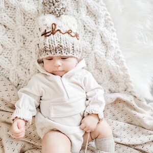 May include: A baby wearing a white knitted hat with a brown pom-pom and the word "three" embroidered on it. The baby is wearing a white onesie and white socks.