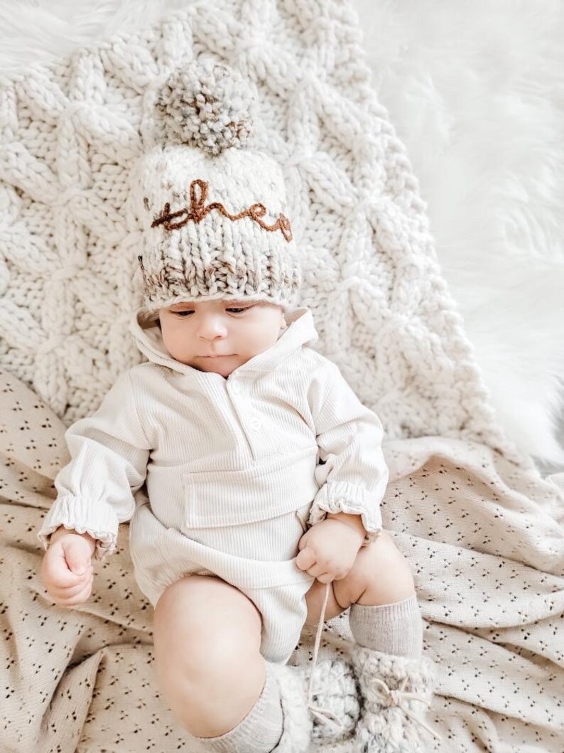 May include: A baby wearing a white knitted hat with a brown pom-pom and the word "three" embroidered on it. The baby is wearing a white onesie and white socks.