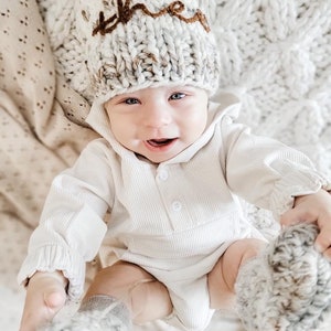 May include: A baby wearing a white knitted hat with the word "the" embroidered in brown yarn. The baby is also wearing a white ribbed onesie and matching knitted booties.