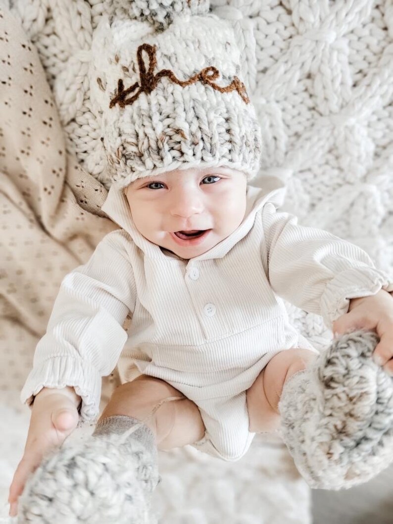 May include: A baby wearing a white knitted hat with the word "the" embroidered in brown yarn. The baby is also wearing a white ribbed onesie and matching knitted booties.